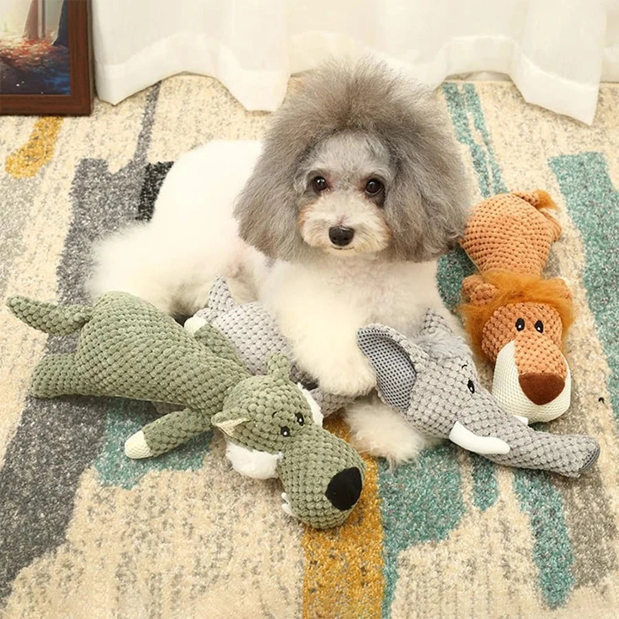 Small dog sitting on a patterned rug with two plush toys, one resembling a wolf and the other a lion.