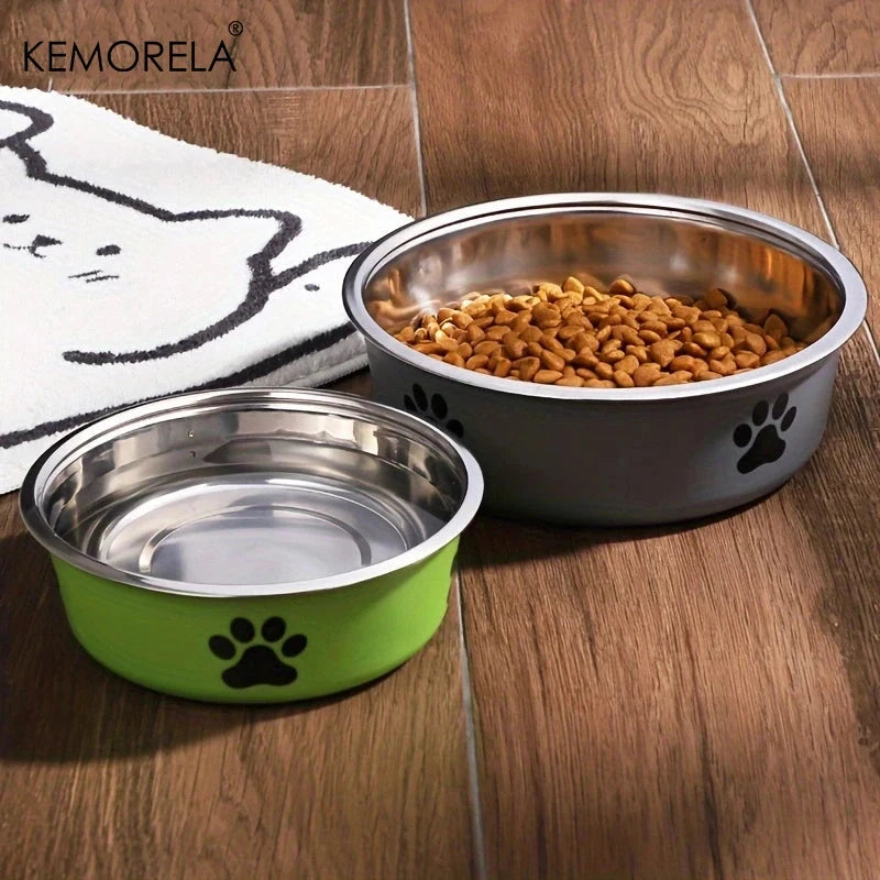 Two stainless steel pet bowls with paw prints, one green and one silver, on a wooden floor with cat food.